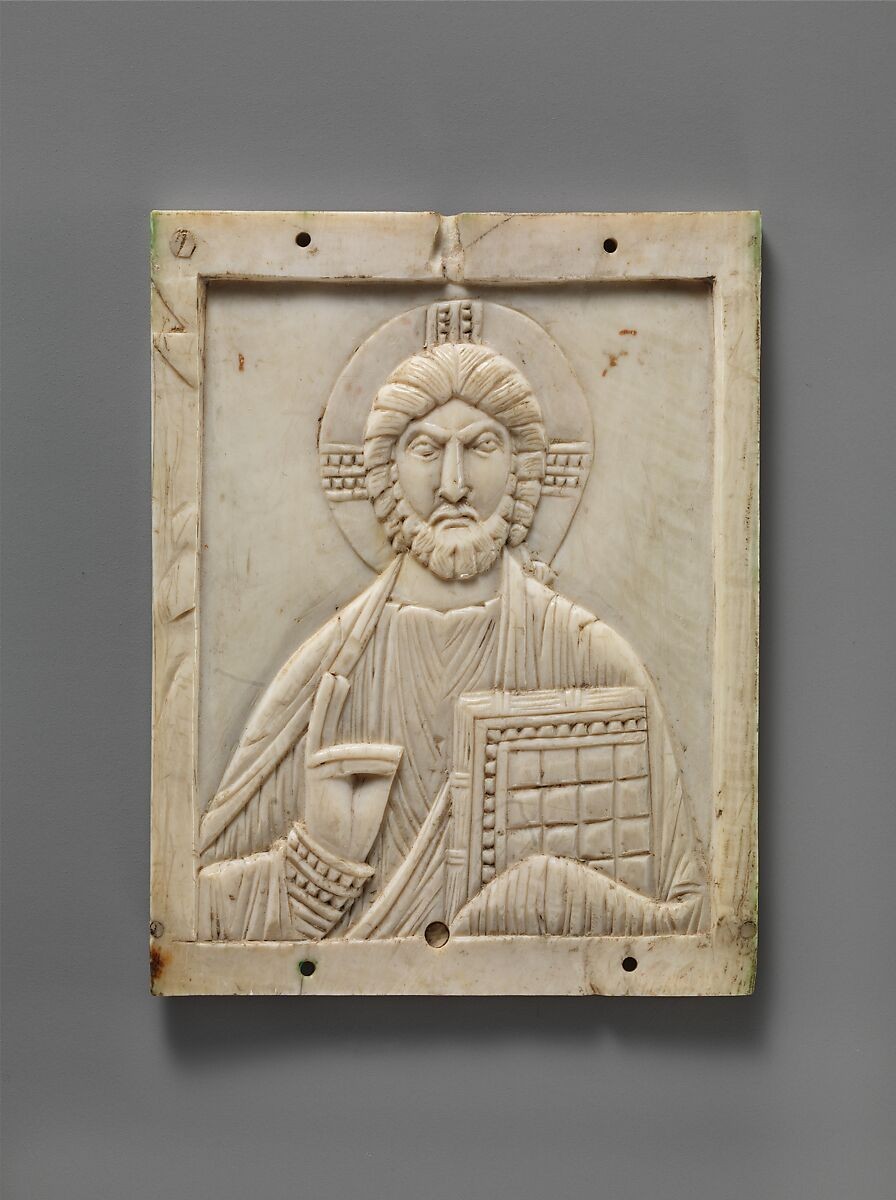 Frontal ivory relief of a haloed Christ, with one hand raised in blessing and the other holding a decorated Gospel book; holes and cracks remain visible.